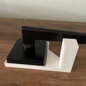 Puede incluir: Una manija de puerta negra con una base blanca. La manija está montada en una base blanca con forma cuadrada. La manija está hecha de metal y tiene un diseño elegante y moderno.