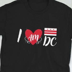 May include: Black t-shirt with the text "I AM DC" in white and red. The design includes a red heart with "AM" inside and a flag graphic. A camera, wallet, phone, and glasses are also visible.