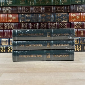 Puede incluir: Un conjunto de tres libros de tapa dura, La Comunidad del Anillo, Las Dos Torres y El Retorno del Rey, de J.R.R. Tolkien. Los libros son verdes con letras doradas y están apilados sobre una superficie de madera.