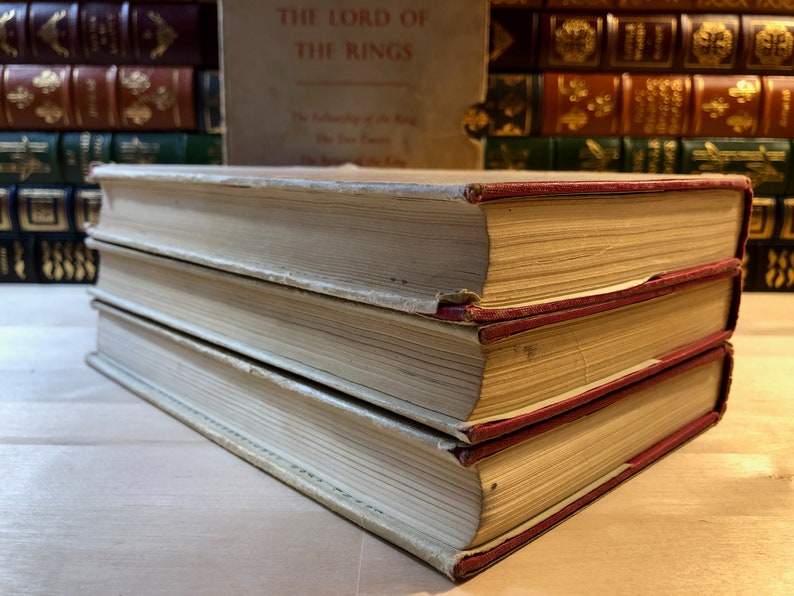 May include: A set of three vintage hardcover books with white covers and red spines. The top book has the title "The Lord of the Rings" printed on the spine.