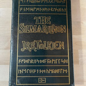 May include: A dark green hardcover book with gold lettering. The title is "The Silmarillion" by J.R.R. Tolkien. The cover features a decorative design of gold symbols.