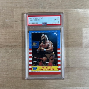 May include: A 1987 Topps WWF Hulk Hogan trading card, graded EX-MT 6, encased in a protective holder. The card features a color image of Hulk Hogan in wrestling attire, with the WWF logo and the text "Hulk Hogan".