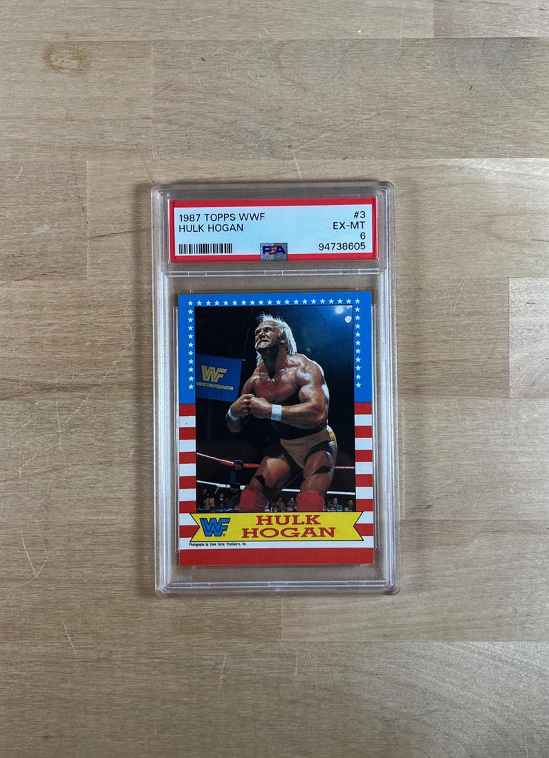 May include: A 1987 Topps WWF Hulk Hogan trading card, graded EX-MT 6, encased in a protective holder. The card features a color image of Hulk Hogan in wrestling attire, with the WWF logo and the text "Hulk Hogan".