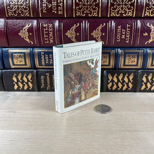 Puede incluir: Un pequeño libro de tapa dura de "Tales of Peter Rabbit" de Beatrix Potter, ilustrado por Charles Santore, se encuentra frente a una pila de libros clásicos. El libro tiene una cubierta blanca con una colorida ilustración de conejos. Una moneda descansa sobre la superficie de madera.