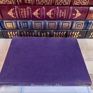 May include: A purple hardcover book titled Pippi Longstocking by Lindgren sits in the foreground. Behind it, several rows of classic books are stacked, featuring burgundy, blue, and black spines with gold embossed lettering and decorative patterns.