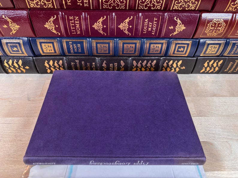 May include: A purple hardcover book titled Pippi Longstocking by Lindgren sits in the foreground. Behind it, several rows of classic books are stacked, featuring burgundy, blue, and black spines with gold embossed lettering and decorative patterns.