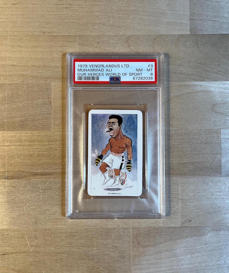 May include: A vintage trading card featuring a cartoon illustration of Muhammad Ali, a famous boxer, in a boxing pose. The card is from the "Our Heroes World of Sport" series and is graded NM-MT 8.