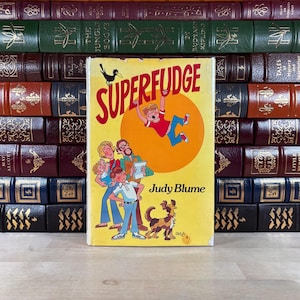 Peut inclure: Un livre jaune intitulé "Superfudge" de Judy Blume, avec une illustration de dessin animé d'un garçon, d'une famille et d'un chien. Le livre est placé devant une collection de livres anciens reliés en cuir.