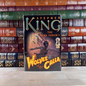 Könnte beinhalten: Ein gebundenes Buch mit dem Titel "The Dark Tower V: Wolves of the Calla" von Stephen King. Das Cover zeigt eine dunkle Illustration einer Person im Rollstuhl. Das Buch steht vor einer Reihe antiker Bücher.