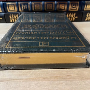 May include: A green hardcover book with gold lettering that reads "J.R.R. TOLKIEN" and "THE FELLOWSHIP OF THE RING" in a stylized font. The book is wrapped in plastic and has a gold-colored spine.