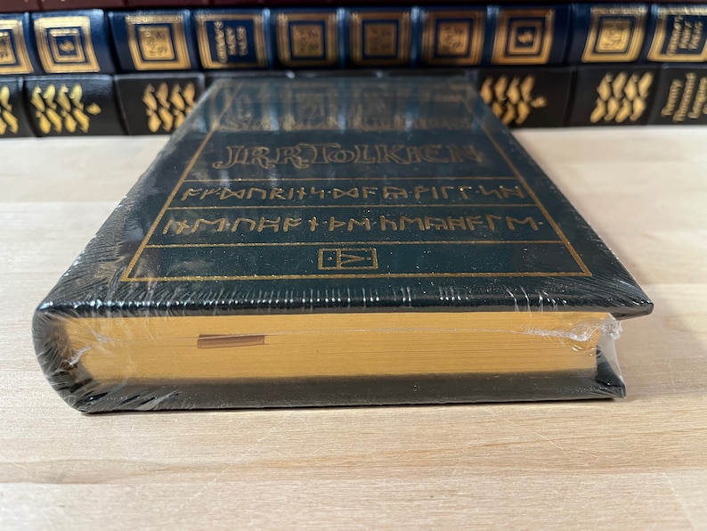 May include: A green hardcover book with gold lettering that reads "J.R.R. TOLKIEN" and "THE FELLOWSHIP OF THE RING" in a stylized font. The book is wrapped in plastic and has a gold-colored spine.
