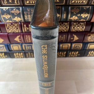 May include: A dark green hardcover book with gold lettering that reads "The Silmarillion" by J.R.R. Tolkien. The book is wrapped in plastic and is sitting on a shelf with other books.