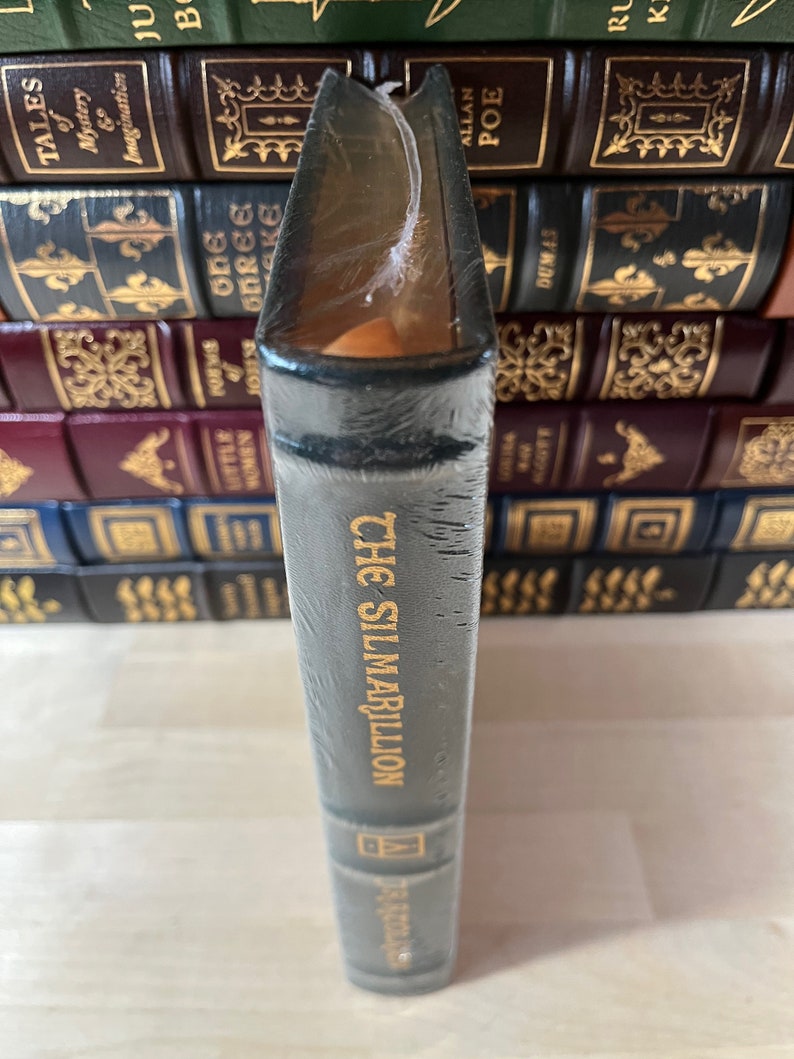 May include: A dark green hardcover book with gold lettering that reads "The Silmarillion" by J.R.R. Tolkien. The book is wrapped in plastic and is sitting on a shelf with other books.