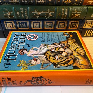 K&ouml;nnte beinhalten: Der Einband des Buches Der hungrige Tiger von Oz zeigt ein M&auml;dchen, das auf einem Tiger reitet, vor einem blauen Hintergrund und mit orangefarbenem R&uuml;cken. Der Titel ist in gro&szlig;en, fetten, wei&szlig;en Buchstaben geschrieben. Das Buch ist von Ruth Plumly Thompson und illustriert von L. Frank Baum.