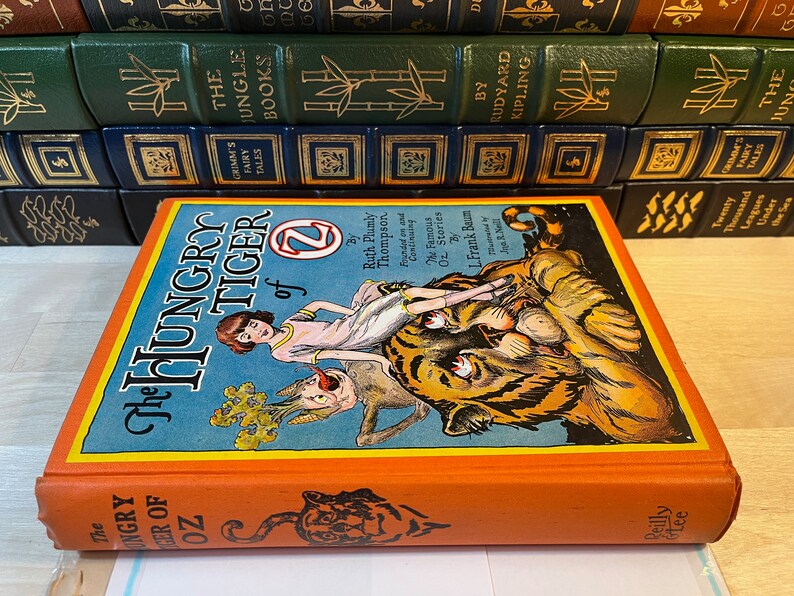 K&ouml;nnte beinhalten: Der Einband des Buches Der hungrige Tiger von Oz zeigt ein M&auml;dchen, das auf einem Tiger reitet, vor einem blauen Hintergrund und mit orangefarbenem R&uuml;cken. Der Titel ist in gro&szlig;en, fetten, wei&szlig;en Buchstaben geschrieben. Das Buch ist von Ruth Plumly Thompson und illustriert von L. Frank Baum.