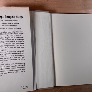 May include: A hardcover book titled Pippi Longstocking by Astrid Lindgren, translated by Florence Lamborn and illustrated by Louis S. Glanzman. The white dust jacket flap features black printed text describing the story and a Book Club Edition label.