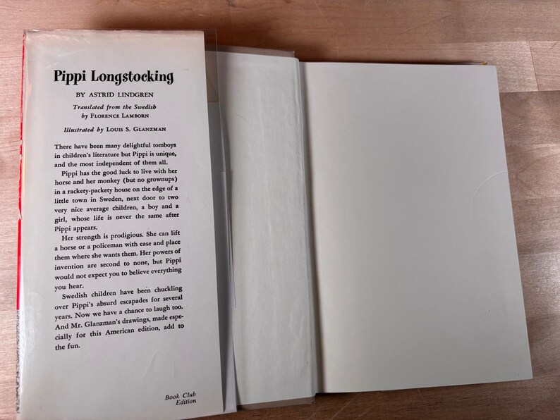 May include: A hardcover book titled Pippi Longstocking by Astrid Lindgren, translated by Florence Lamborn and illustrated by Louis S. Glanzman. The white dust jacket flap features black printed text describing the story and a Book Club Edition label.