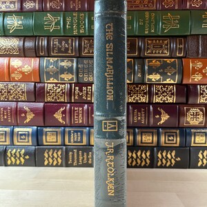 May include: A dark green hardcover book with gold lettering. The title is "The Silmarillion" by J.R.R. Tolkien. The book is on a wooden surface.