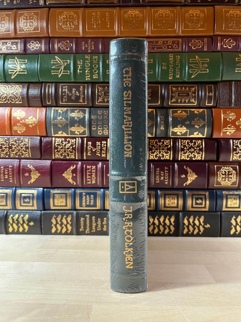 May include: A dark green hardcover book with gold lettering. The title is "The Silmarillion" by J.R.R. Tolkien. The book is on a wooden surface.
