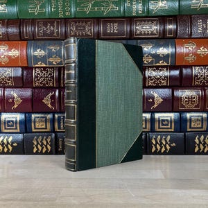 May include: A vintage book with a green cover and gold accents stands in front of a collection of antique books. The spine is dark green with gold lettering. The book's cover is a textured green with a gold border.