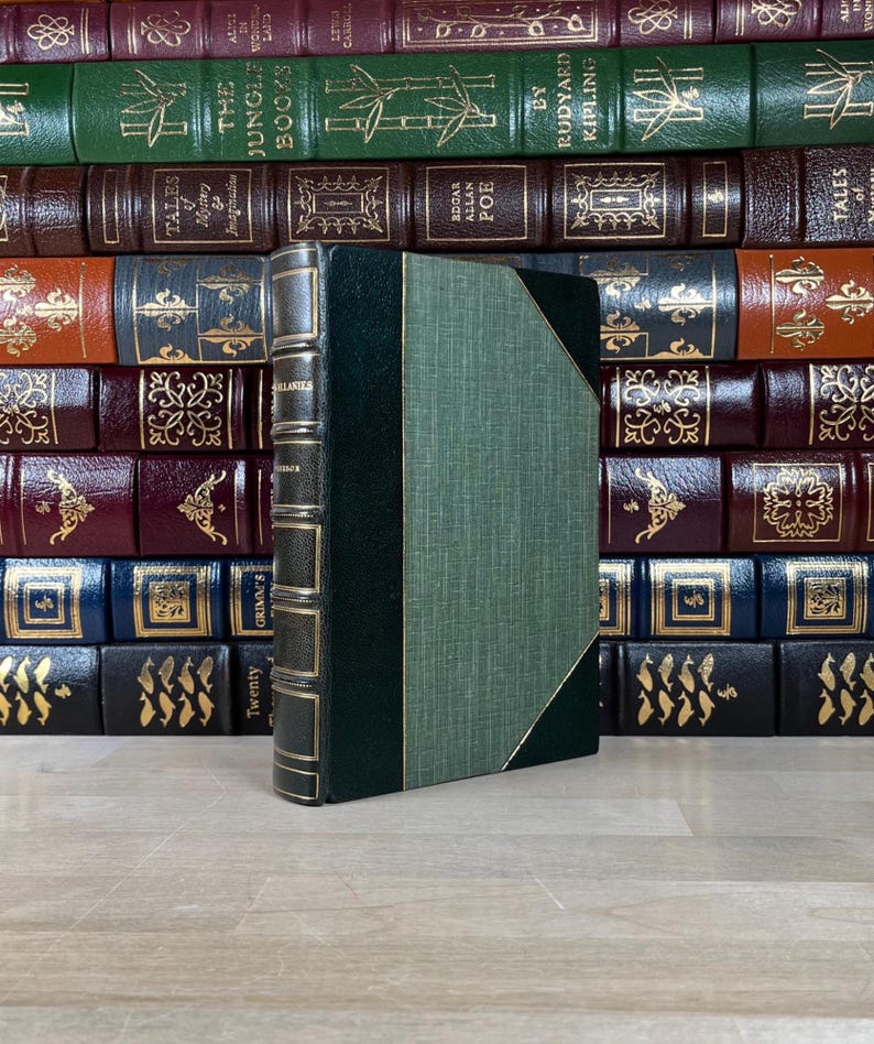 May include: A vintage book with a green cover and gold accents stands in front of a collection of antique books. The spine is dark green with gold lettering. The book's cover is a textured green with a gold border.