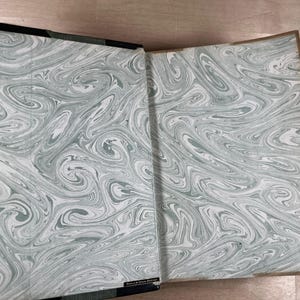 May include: Open book with marbled endpapers in shades of gray and white. The book's spine is black with a green edge. The marbled pattern creates a swirling, organic design. The book is open, revealing the inside cover.