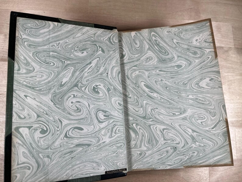 May include: Open book with marbled endpapers in shades of gray and white. The book's spine is black with a green edge. The marbled pattern creates a swirling, organic design. The book is open, revealing the inside cover.