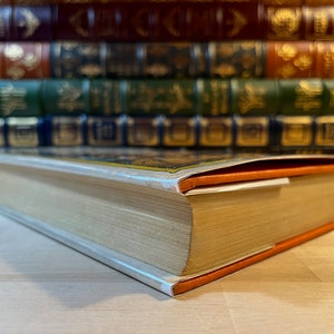 K&ouml;nnte beinhalten: Nahaufnahme des Buchr&uuml;ckens eines gebundenen Buches mit einem orangefarbenen und wei&szlig;en Schutzumschlag. Das Buch liegt auf einer Holzoberfl&auml;che.