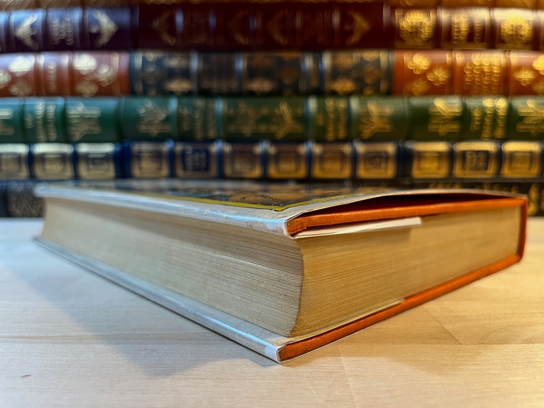 K&ouml;nnte beinhalten: Nahaufnahme des Buchr&uuml;ckens eines gebundenen Buches mit einem orangefarbenen und wei&szlig;en Schutzumschlag. Das Buch liegt auf einer Holzoberfl&auml;che.