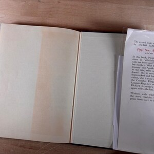 May include: An open hardcover book with cream-colored pages and a white dust jacket flap. The text on the flap reads: The second book about Pippi by ASTRID LINDGREN is Pippi Goes Aboard. In this book, Pippi still lives alone in Villekulla cottage.