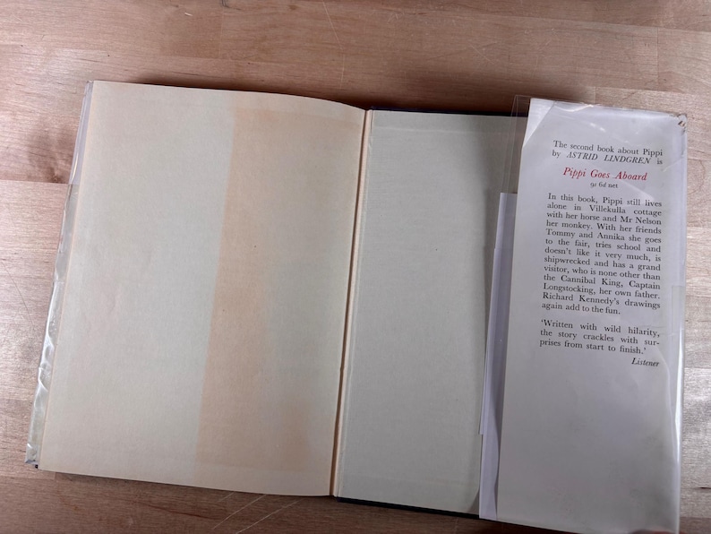 May include: An open hardcover book with cream-colored pages and a white dust jacket flap. The text on the flap reads: The second book about Pippi by ASTRID LINDGREN is Pippi Goes Aboard. In this book, Pippi still lives alone in Villekulla cottage.