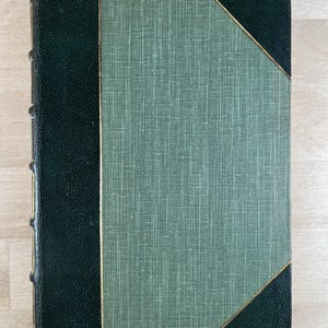 May include: A vintage book with a textured green cover and dark green leather spine and corners. The cover features a diagonal gold trim. The book's spine has gold detailing.