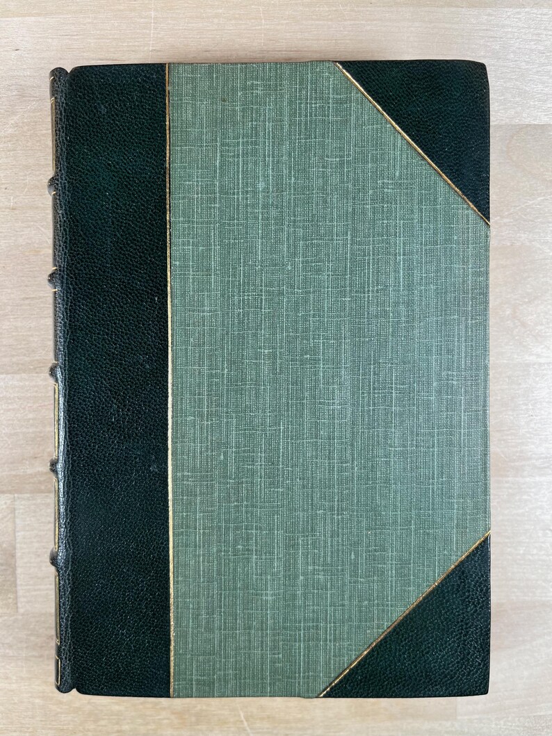 May include: A vintage book with a textured green cover and dark green leather spine and corners. The cover features a diagonal gold trim. The book's spine has gold detailing.