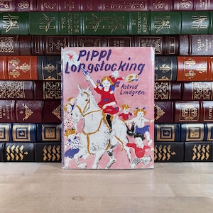 Puede incluir: Un ejemplar vintage de "Pippi Longstocking" de Astrid Lindgren, con una portada rosa que muestra a Pippi a caballo. El libro se presenta frente a una colecci&oacute;n de libros antiguos encuadernados en cuero de varios colores.