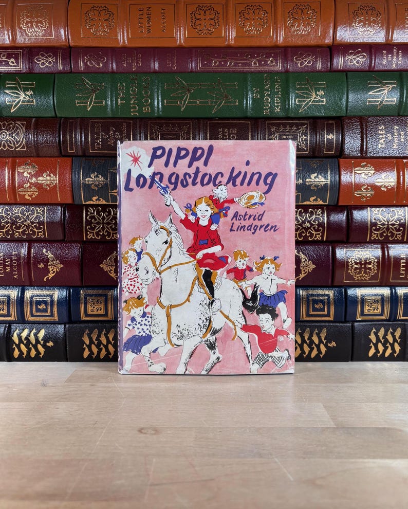 Puede incluir: Un ejemplar vintage de "Pippi Longstocking" de Astrid Lindgren, con una portada rosa que muestra a Pippi a caballo. El libro se presenta frente a una colecci&oacute;n de libros antiguos encuadernados en cuero de varios colores.