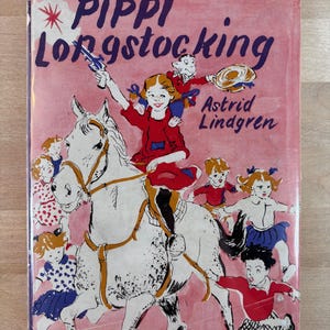 Puede incluir: Portada de libro infantil vintage con el t&iacute;tulo "Pippi Longstocking" en letras moradas sobre un fondo rosa. La ilustraci&oacute;n muestra a una ni&ntilde;a montada en un caballo blanco, rodeada de otros ni&ntilde;os. Tambi&eacute;n se ve el nombre de la autora, Astrid Lindgren.