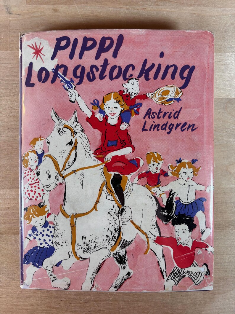 Puede incluir: Portada de libro infantil vintage con el t&iacute;tulo "Pippi Longstocking" en letras moradas sobre un fondo rosa. La ilustraci&oacute;n muestra a una ni&ntilde;a montada en un caballo blanco, rodeada de otros ni&ntilde;os. Tambi&eacute;n se ve el nombre de la autora, Astrid Lindgren.