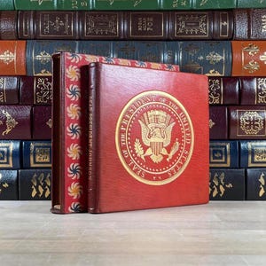 May include: A stack of antique books with leather covers. The front book is red with a gold seal and the text "SEAL OF THE PRESIDENT OF THE UNITED STATES". The spine reads "DEAR PRESIDENT JOHNSON".