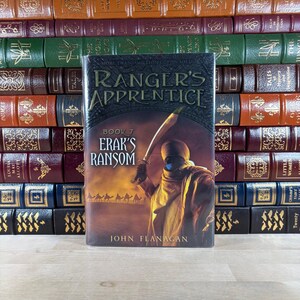 May include: A book titled "Ranger's Apprentice, Book 7: Erak's Ransom" by John Flanagan. The cover features a warrior with a sword and a desert scene. The book is set against a backdrop of antique books.