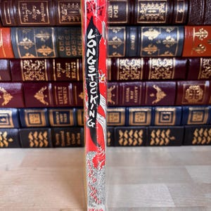 May include: A book spine with a red and yellow background featuring the black text LONGSTOCKING vertically. The author name ASTRID LINDGREN is printed in black at the bottom, with the word VIKING below it. The book is encased in clear plastic.