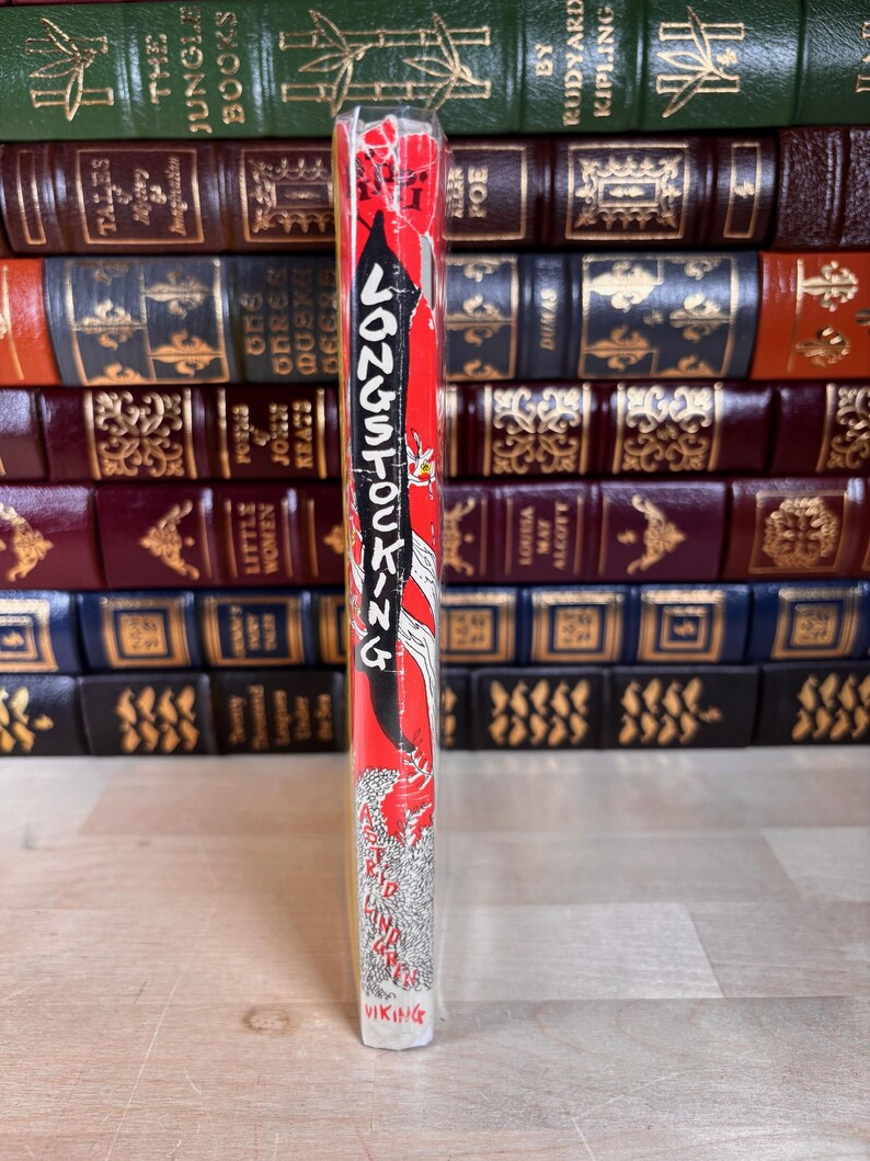 May include: A book spine with a red and yellow background featuring the black text LONGSTOCKING vertically. The author name ASTRID LINDGREN is printed in black at the bottom, with the word VIKING below it. The book is encased in clear plastic.