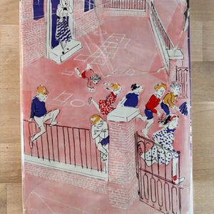 May include: A rectangular book cover featuring a pink-toned illustration of children playing in a courtyard. The scene depicts a person standing in a doorway, children climbing a metal fence, and others running near a gate and hopscotch grid.