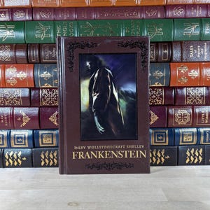 May include: A hardcover edition of Mary Wollstonecraft Shelley's "Frankenstein" with a dark brown cover. The book features an illustration of a figure in a coat and the title in gold lettering. A stack of antique books forms the background.