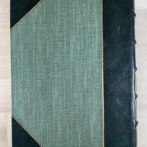May include: A vintage book with a textured green cover and dark green leather spine and corners. The cover features a diagonal gold trim. The spine has gold accents. The book is closed and in good condition.