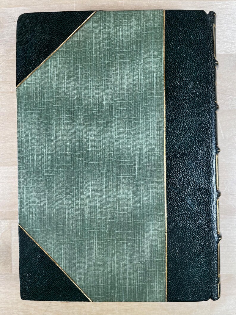 May include: A vintage book with a textured green cover and dark green leather spine and corners. The cover features a diagonal gold trim. The spine has gold accents. The book is closed and in good condition.