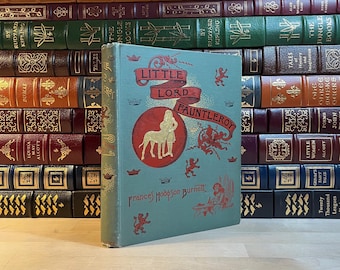 Może przedstawiać: Antyczna książka w twardej oprawie zatytułowana "Mały Lord Fauntleroy" autorstwa Frances Hodgson Burnett. Okładka książki przedstawia zielone tło z czerwonym kołem, w którym znajduje się biały pies i dziewczynka w niebieskiej sukience. Książka jest otoczona innymi książkami na drewnianej powierzchni.