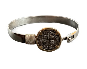 May include: A silver bangle bracelet with a gold-toned coin charm featuring a cross design. The bracelet has a hinged clasp.