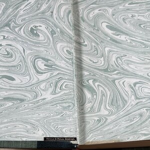 May include: Open book with marbled endpapers. The design features swirling patterns in shades of gray and white, creating a flowing, abstract aesthetic. The book's spine and edges are visible, adding context to the design.