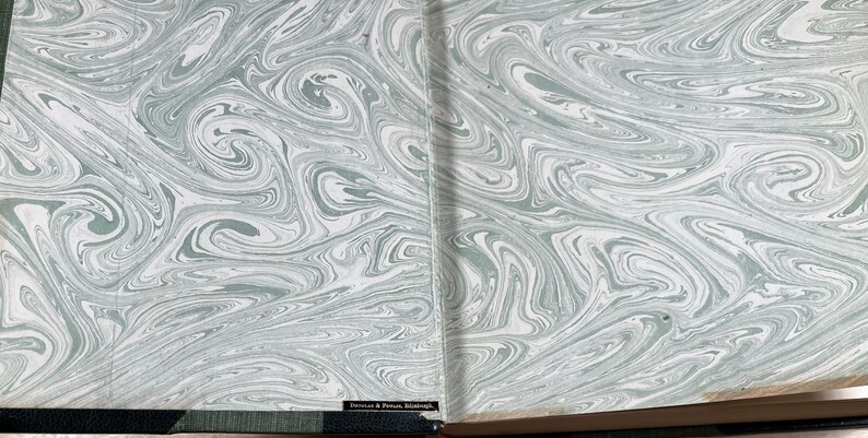 May include: Open book with marbled endpapers. The design features swirling patterns in shades of gray and white, creating a flowing, abstract aesthetic. The book's spine and edges are visible, adding context to the design.