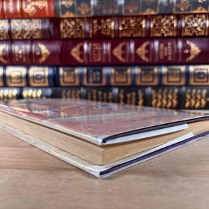 May include: A single paperback book with a reddish-brown cover lies flat in the foreground. Behind it, a row of five hardcover books with ornate, gold-embossed spines in shades of red, burgundy, and dark blue are arranged horizontally on a shelf.